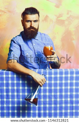 alcohol cocktail holding flask or bottle on blue checkered plaid on colorful texture background. Bearded man, long beard. Brutal caucasian tattooed serious hipster with moustache in denim shirt with