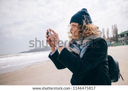 Girl tourist taking a picture of ocean shore using smart phone at winter day. Young woman with super curly red hair wearing hat, scarf and coat making photo of seascape.
