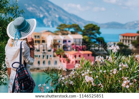 Blond beautiful woman in blue sun hat and white clothes enjoying view of colorful tranquil village Assos houses on sunny day. Stylish female visiting Kefalonia in summer time on Greece travel vacation Royalty-Free Stock Photo #1146525851