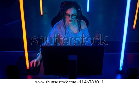 High Angle Shot of the Beautiful Friendly Pro Gamer Girl Playing in Online Video Game and Streaming it, Wearing Headset Talks with Her Fans into Headphones Microphone. Background Cool Neon Retro Color