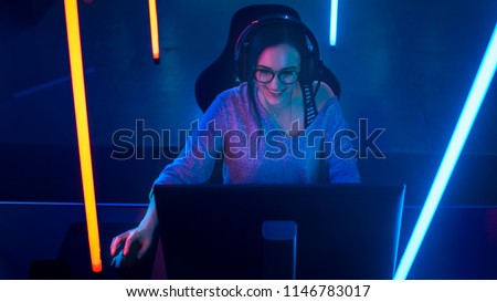 High Angle Shot of the Beautiful Friendly Pro Gamer Girl Playing in Online Video Game and Streaming it, Wearing Headset Talks with Her Fans into Headphones Microphone. Background Cool Neon Retro Color