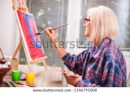 Elderly woman is painting in her home. Retirement hobby. 