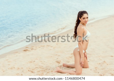 attractive brunette girl posing in white bikini on sandy beach at sea