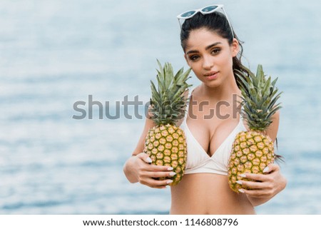 girl in white bikini holding sweet pineapples near the sea