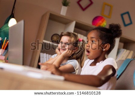 Two beautiful little girls sitting at a desk, watching cartoons on a laptop computer and having fun