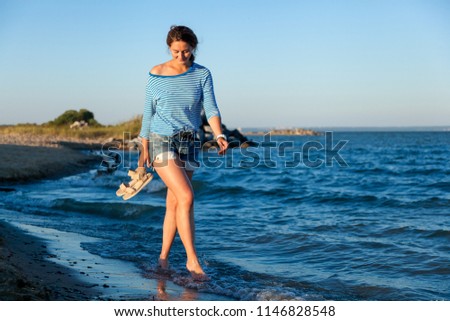 A cheerful dark-haired woman in jeans and a striped T-shirt smiles, walks along the beach and enjoys the bright sun on a summer day. Concept of summer holidays at sea and live style
