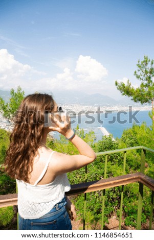 Young beautiful travelling tourist girl woman taking photo shot of resort city and sea coast line, typing at smart phone, searching for cellular data signal, summer vacation concept, sunny outdoors 