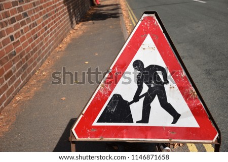 Old, weathered, red and white triangular road safety sign Men At Work on the side of the road. Black symbol person digging on white background