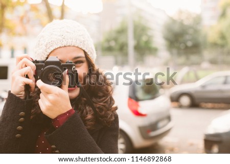 Young beautiful woman with a woolen cap taking pictures with a retro camera in the city in autumn. Focus on camera