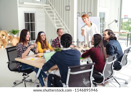 Group of multiracial young creative team talking, laughing and brainstorming in meeting at modern office concept. Female standing gesture hand for sharing while others sitting together in rear view.