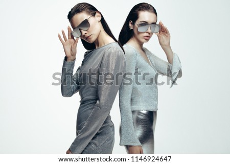 Portrait of two young females in silver outfit and mirrored sunglasses looking at camera. Female models in futuristic look standing on grey background.