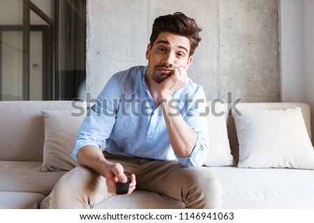 Bored young man holding tv remote control while sitting on a couch at home Royalty-Free Stock Photo #1146941063