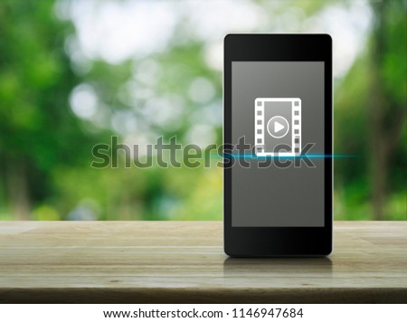 Play button with movie icon on modern smart mobile phone screen on wooden table over blur green tree in park, Business cinema online concept