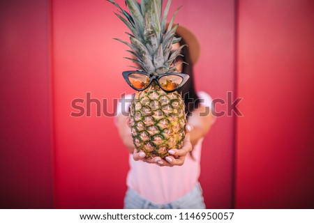 A picture of girl holds pineapple. She shows in on camera. It has glasses on. Isolated on red and striped background.