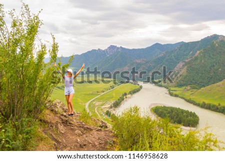 A young blond woman in a white cap, blue denim shorts and a striped T-shirt is standing on a rock by the river of the katun in the Altai mountains raising her hands upwards