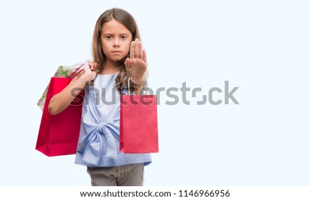 Brunette hispanic girl holding shopping bags with open hand doing stop sign with serious and confident expression, defense gesture
