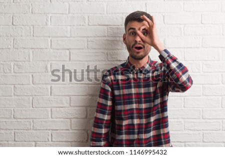 Young adult man standing over white brick wall doing ok gesture shocked with surprised face, eye looking through fingers. Unbelieving expression.