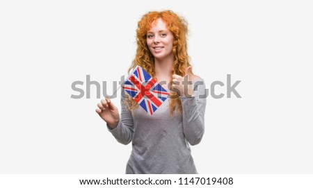 Young redhead woman holding flag of United Kingdom happy with big smile doing ok sign, thumb up with fingers, excellent sign