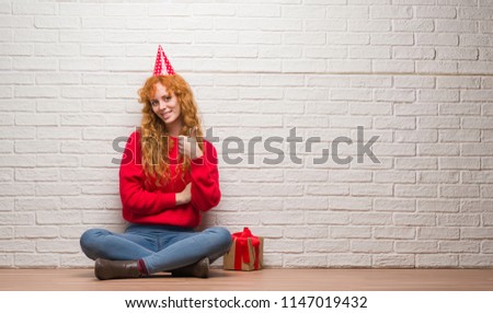 Young redhead woman sitting over brick wall wearing birthday hat happy with big smile doing ok sign, thumb up with fingers, excellent sign