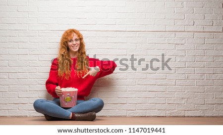 Young redhead woman sitting over brick wall eating popcorn very happy pointing with hand and finger