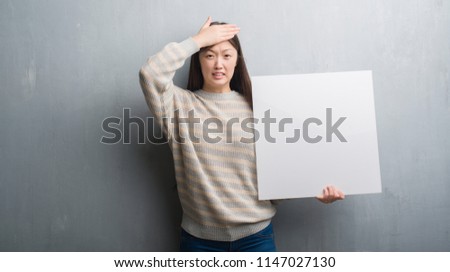 Young Chinese woman over grey wall holding banner stressed with hand on head, shocked with shame and surprise face, angry and frustrated. Fear and upset for mistake.