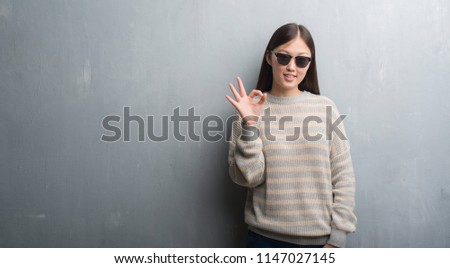 Young Chinese woman over grey wall wearing sunglasses doing ok sign with fingers, excellent symbol