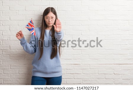 Young Chinese woman over brick wall holding flag of England with open hand doing stop sign with serious and confident expression, defense gesture