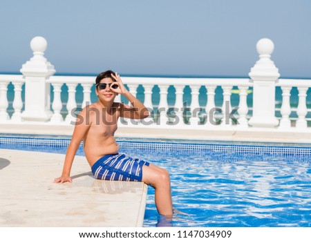 Young child on holiday at the swimming pool by the beach with happy face smiling doing ok sign with hand on eye looking through fingers