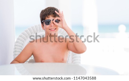 Young child on holidays sitting on a chair by the beach with happy face smiling doing ok sign with hand on eye looking through fingers