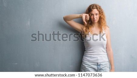 Young redhead woman over grey grunge wall with angry face, negative sign showing dislike with thumbs down, rejection concept