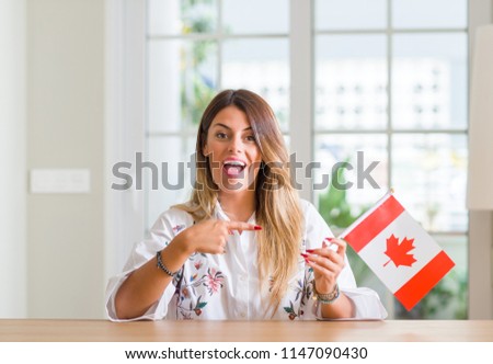 Young woman at home holding flag of Canada very happy pointing with hand and finger