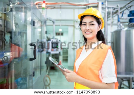 Young engineer inspecting production line. Young beautiful chinese woman in safety hat holding tablet with bottle filling production line in background. Royalty-Free Stock Photo #1147098890