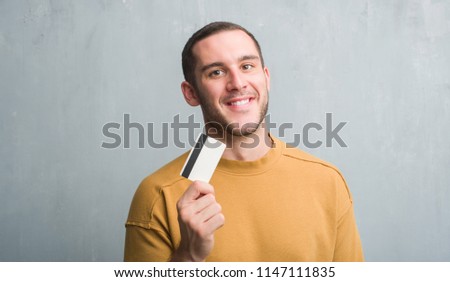 Young caucasian man over grey grunge wall holding credit card with a happy face standing and smiling with a confident smile showing teeth