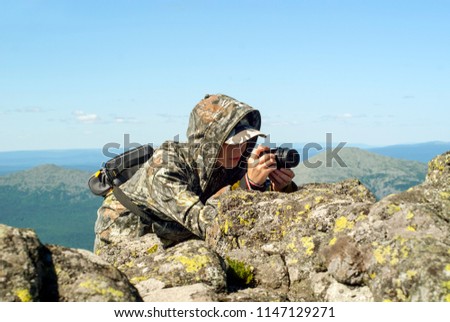 girl wildlife photographer in camouflage suit in the mountains hiding behind a stone photographing something