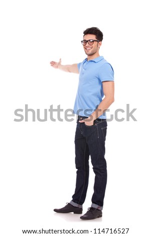 Full body picture of a happy young casual man presenting something in the back with a hand in pocket and looking at the camera