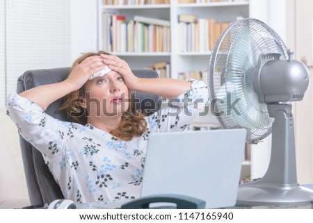 Woman suffers from heat while working in the office and tries to cool off by the fan Royalty-Free Stock Photo #1147165940