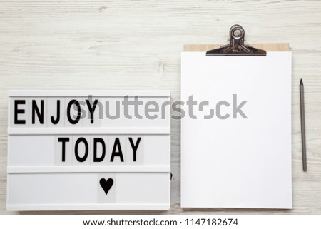 Work space with noticepad, pencil and 'Enjoy today' word on lightbox over white wooden background, top view. From above, flat-lay, overhead. 