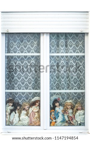 Closeup view of a white window with modern shuttter, old curtain, several vintage beautiful caucasian porcelain dolls and a clown,  behind the pane, looking in the same direction, France.