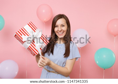 Portrait of pretty smiling woman in blue dress untying bow on red box with gift present on pastel pink background with colorful air balloons. Birthday holiday party, people sincere emotions concept