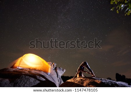 Attractive athletic girl doing complicated gymnastic exercises on big boulder on starry sky and brightly lit tourist tent background. Tourism, hiking, active lifestyle, mountain night camping concept.