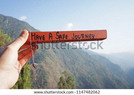 Close up hand holding wooden guest house key chain with text 'HAVE A SAFE JOURNEY ' in natural landscape of green mountain range