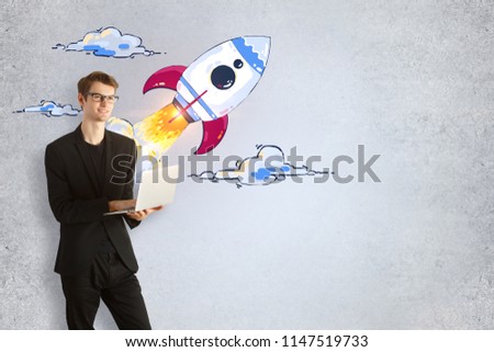 Businessman with laptop and creative launching rocket sketch on concrete wall background. Startup and technology concept