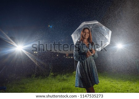 Woman in a dress in the rain with a transparent umbrella in the night. Smiling young woman in the rain with an umbrella. Beautiful woman with a transparent umbrella in the lanterns and rain drop