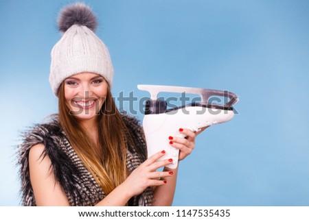 Winter sport activity concept. Woman wearing furry warm hat holding ice skate, blue background studio shot.
