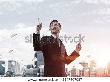 Horizontal shot of cheerful and young businessman in black suit gesturing and smiling while standing against cityscape view with flying paper planes and cloudy sky on background.