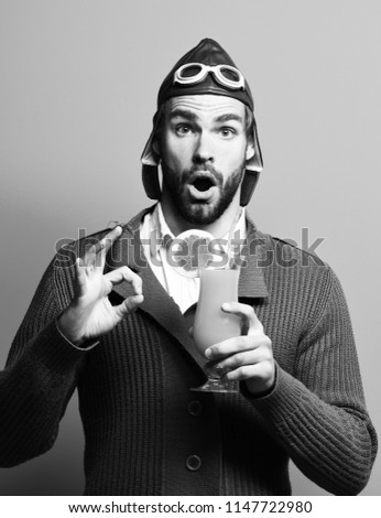handsome bearded aviator man with long beard on surprised face holding glass of alcoholic cocktail in gray knitted sweater with hat and glasses on blue studio background.