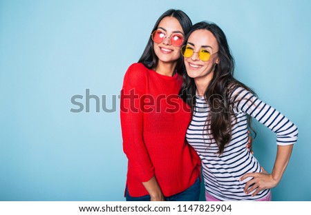 Two beautiful brunette women in sunglasses posing and hugging over blue background