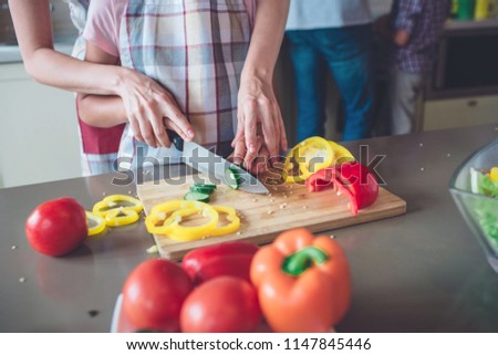 A picture of colorful and tasty food lying on the table. There are peppers, tomatoes cucumber. Mother hepls her daughter to cut green vegetable.