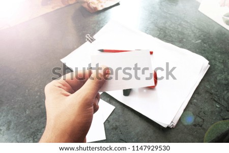 Hand holding a plain white business card with papers,stamp and a red pen in the background.