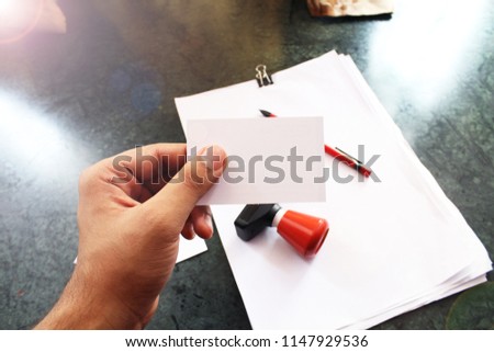 Hand holding a plain white business card with papers,stamp and a red pen in the background.